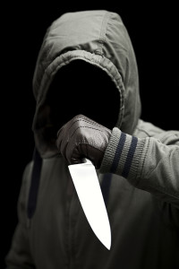 A terrifying mysterious man wearing a hooded jacket with a sharp knife in a striking pose. Shot against a black background.